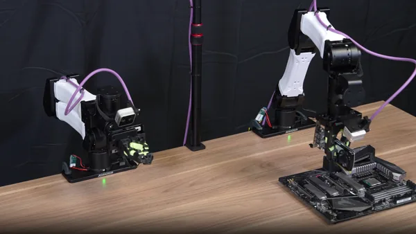 Two robotic arms sit on a lab table, with the robotic arm on the right holding a GPU above a motherboard. Teams of AI coding agents can train robots to do various manipulation tasks.